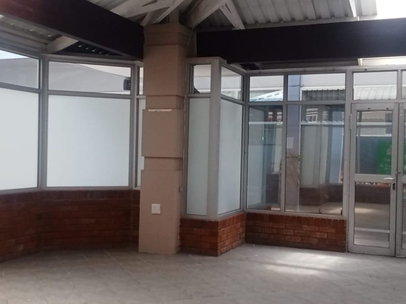 A bright, empty retail entrance area featuring exposed brickwork, large aluminium-framed glass windows, and tiled flooring, suitable for a professional storefront or reception space.