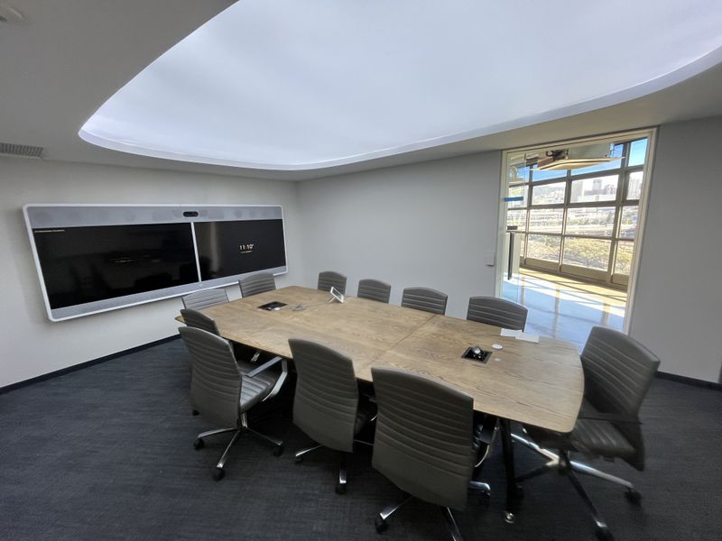 Modern professional boardroom featuring a large conference table, integrated wall-mounted display screen, recessed ceiling lighting, and expansive windows with natural light.