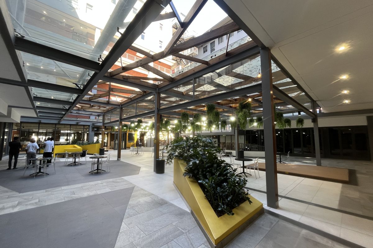A modern, covered outdoor atrium featuring a glass roof, steel structural framing, and integrated planters, providing a bright and versatile communal space.