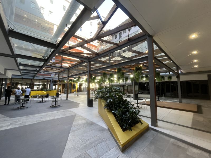 A modern, covered outdoor atrium featuring a glass roof, steel structural framing, and integrated planters, providing a bright and versatile communal space.