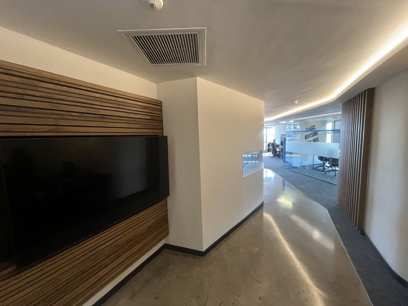 Modern office corridor featuring polished concrete floors, timber slat wall accents, recessed LED lighting, and glass partitions leading to an open-plan workspace.