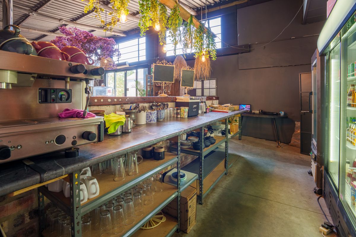 A functional commercial kitchen area featuring industrial shelving, a service counter, and a commercial refrigerator within an open-plan restaurant space.