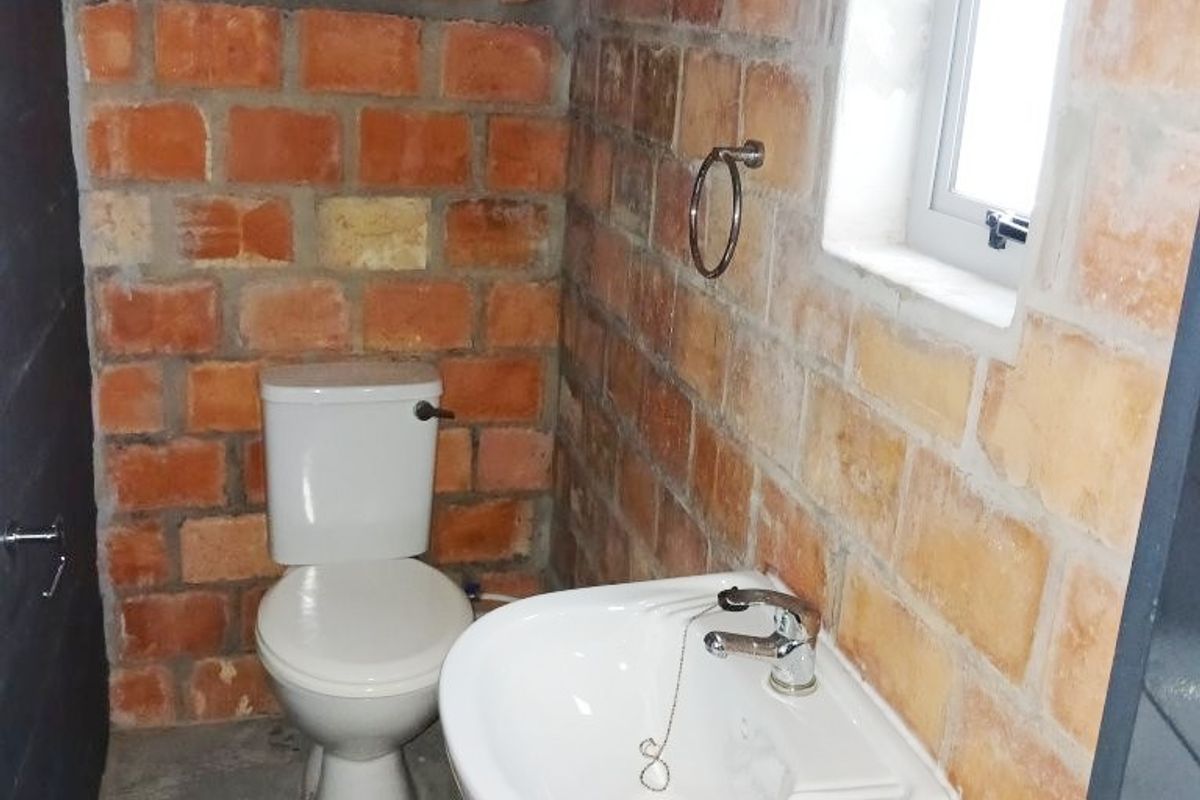A compact bathroom featuring exposed brick walls, a wall-mounted basin, and a standard toilet, currently in an unfinished, industrial-style condition.