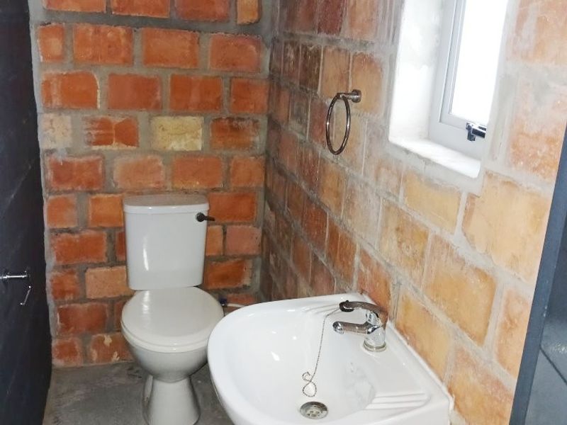 A compact bathroom featuring exposed brick walls, a wall-mounted basin, and a standard toilet, currently in an unfinished, industrial-style condition.