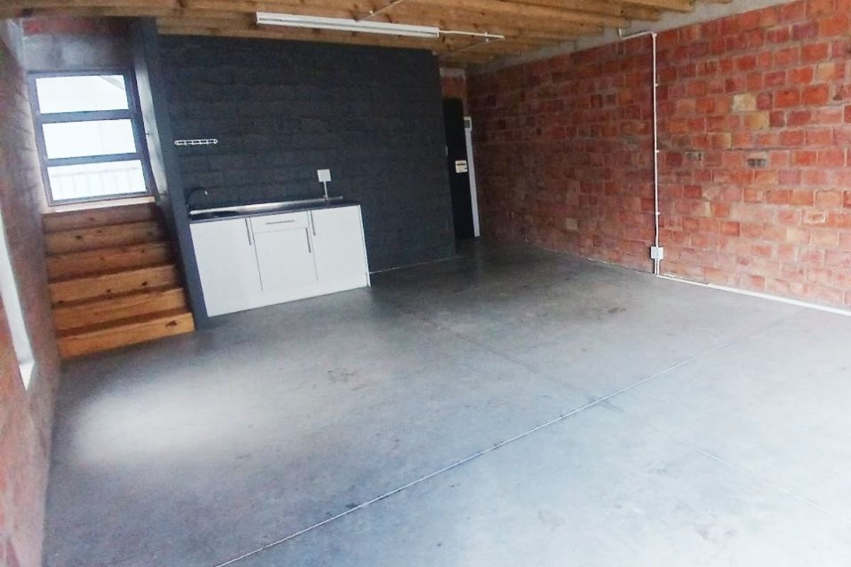 An open-plan office space featuring exposed brick walls, timber ceiling beams, polished concrete floors, and a compact kitchenette area with a sink.