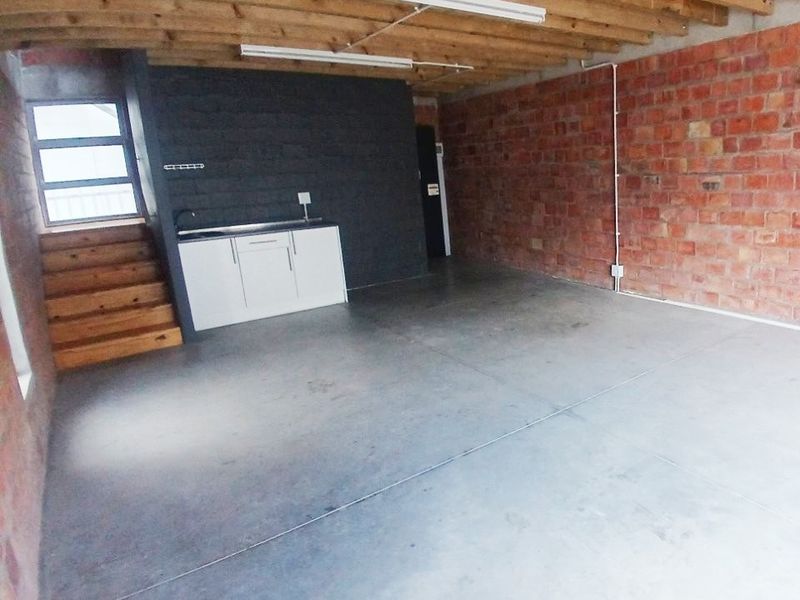 An open-plan office space featuring exposed brick walls, timber ceiling beams, polished concrete floors, and a compact kitchenette area with a sink.
