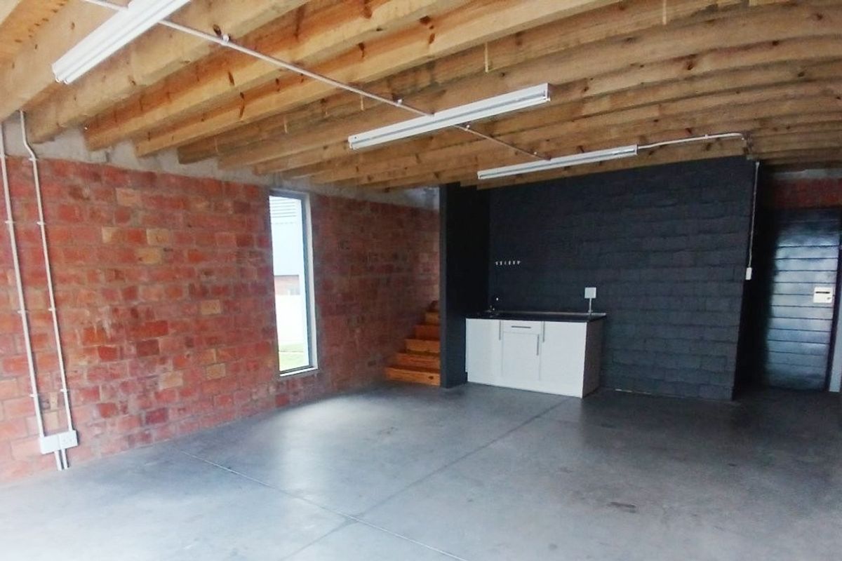 An open-plan office space featuring exposed brick walls, timber ceiling joists, polished concrete floors, and a compact kitchenette area.