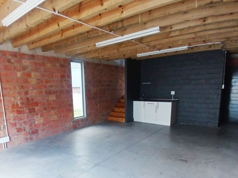 An open-plan office space featuring exposed brick walls, timber ceiling joists, polished concrete floors, and a compact kitchenette area.