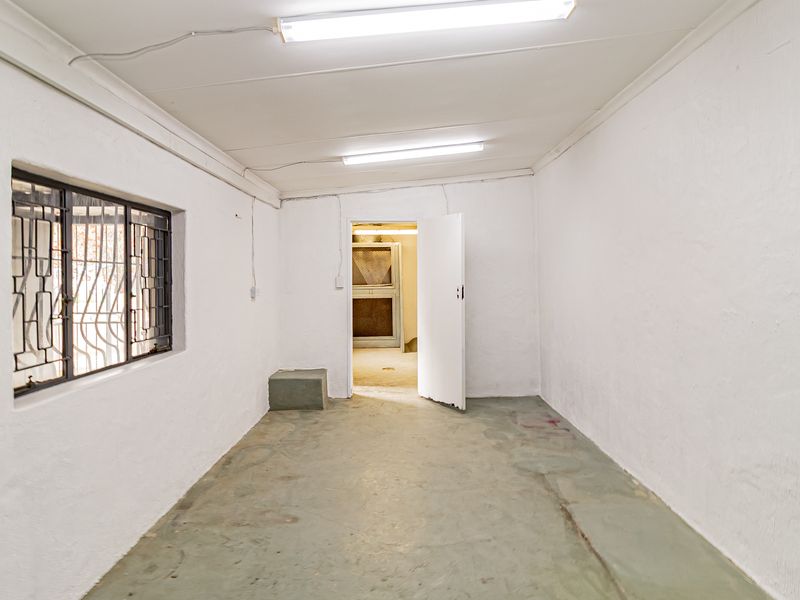 A vacant office space featuring white painted walls, concrete flooring, security bars on the window, and overhead fluorescent lighting.