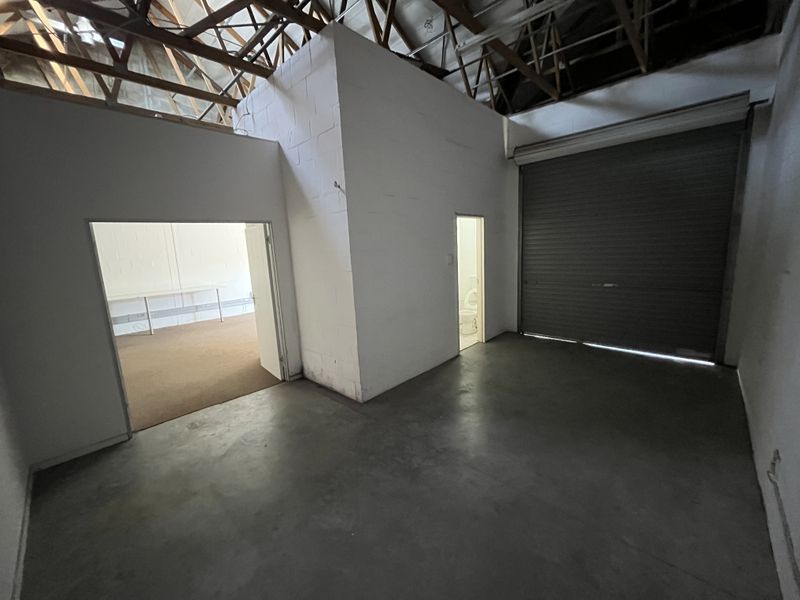 Industrial utility space featuring concrete flooring, exposed timber roof trusses, and a large roller shutter door, with access to an adjoining office and restroom.