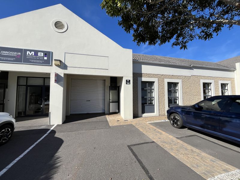 Front elevation of an industrial unit featuring a roller shutter door, paved parking area, and professional signage.