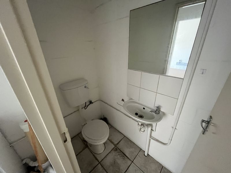 A compact industrial-style bathroom featuring a wall-mounted basin, toilet, and tiled flooring, requiring cosmetic updates to improve its overall condition.