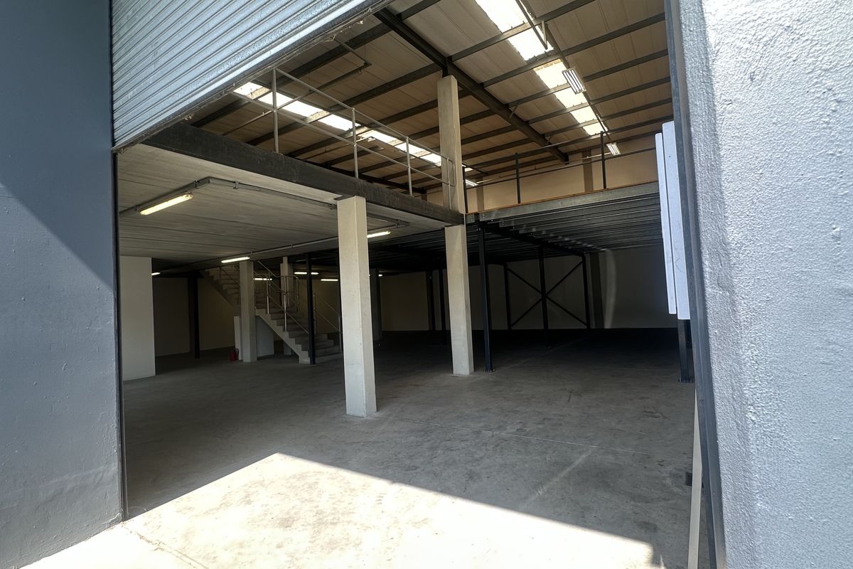 Spacious industrial warehouse unit featuring a large roller shutter door, concrete flooring, and a functional mezzanine level with integrated staircase and overhead lighting.