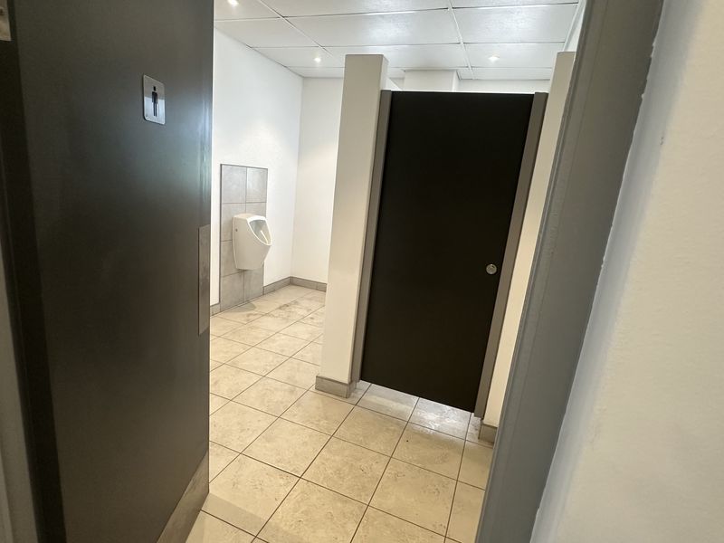 A functional industrial-style restroom featuring a wall-mounted urinal, a private toilet cubicle, tiled flooring, and a suspended ceiling with recessed lighting.