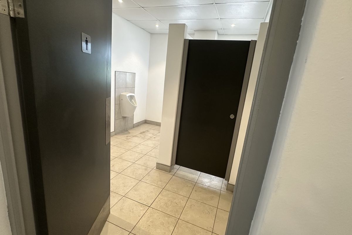 A functional industrial-style restroom featuring a wall-mounted urinal, a private toilet cubicle, tiled flooring, and a suspended ceiling with recessed lighting.