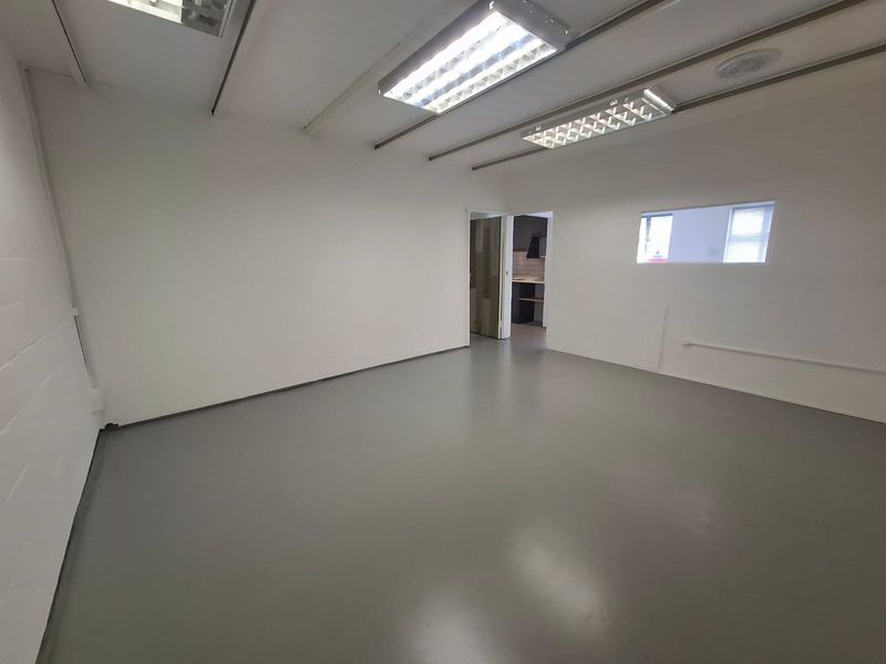 A clean, vacant industrial office space featuring durable epoxy flooring, bright fluorescent lighting, and an internal window looking into an adjacent area.