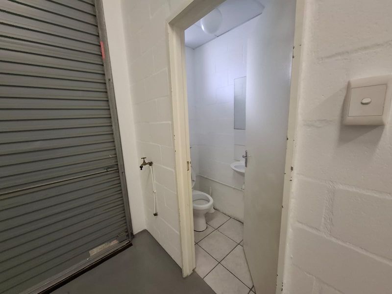 A functional industrial-style bathroom featuring a toilet and wall-mounted basin, accessed via a doorway adjacent to a roller shutter door.