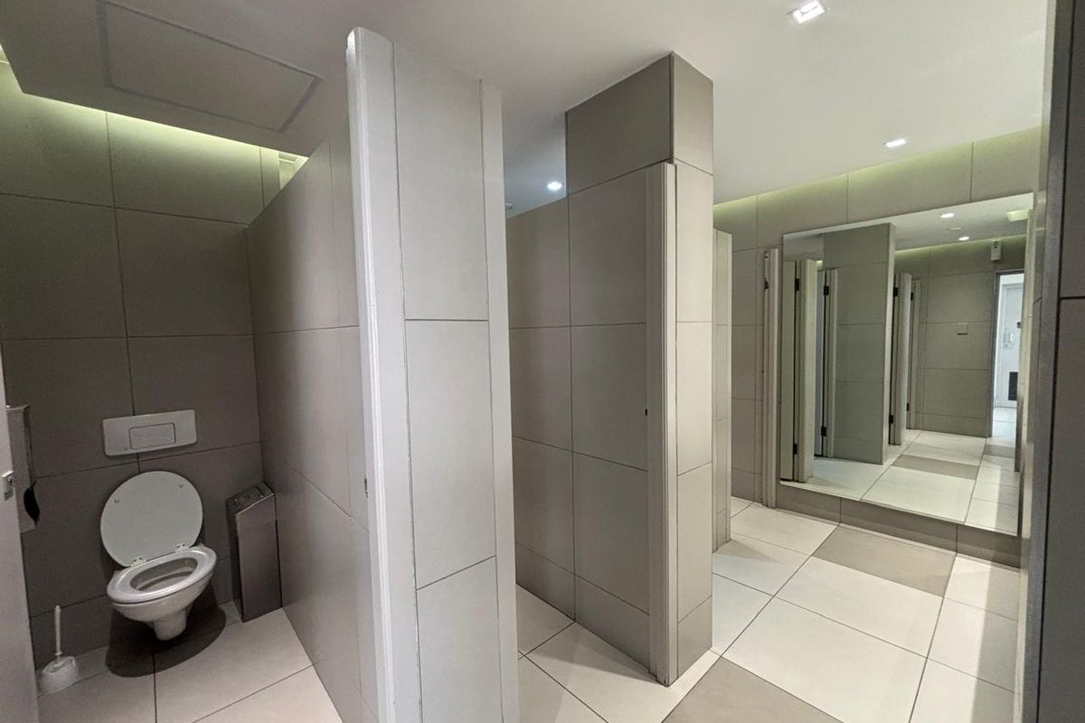 Modern office bathroom featuring tiled partitions, wall-mounted toilets with concealed cisterns, and a large vanity mirror with recessed lighting.