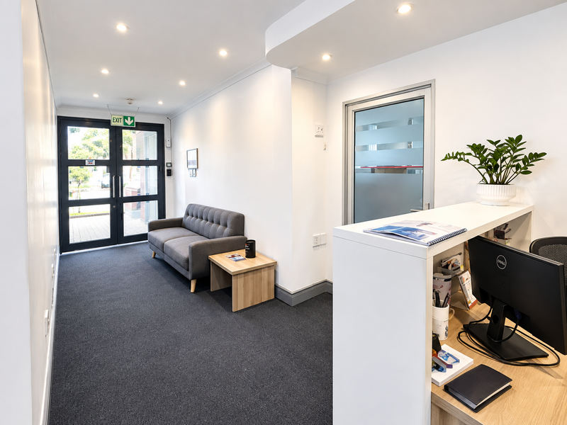 A professional office reception area featuring a built-in desk, recessed lighting, carpeted flooring, and a glass-paneled entrance door.