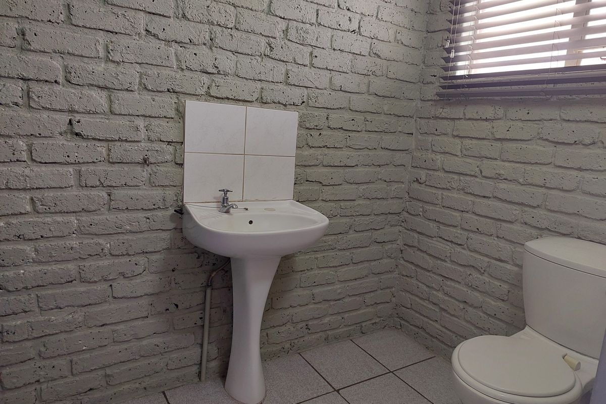 A functional bathroom featuring a pedestal sink, toilet, tiled flooring, and painted brick walls with window blinds.