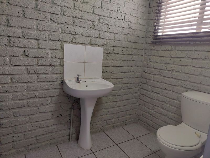 A functional bathroom featuring a pedestal sink, toilet, tiled flooring, and painted brick walls with window blinds.