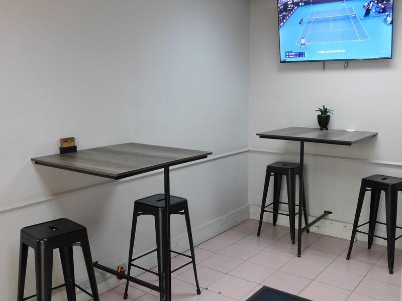 A functional office break area featuring wall-mounted high tables, tiled flooring, and wall-mounted television screen.