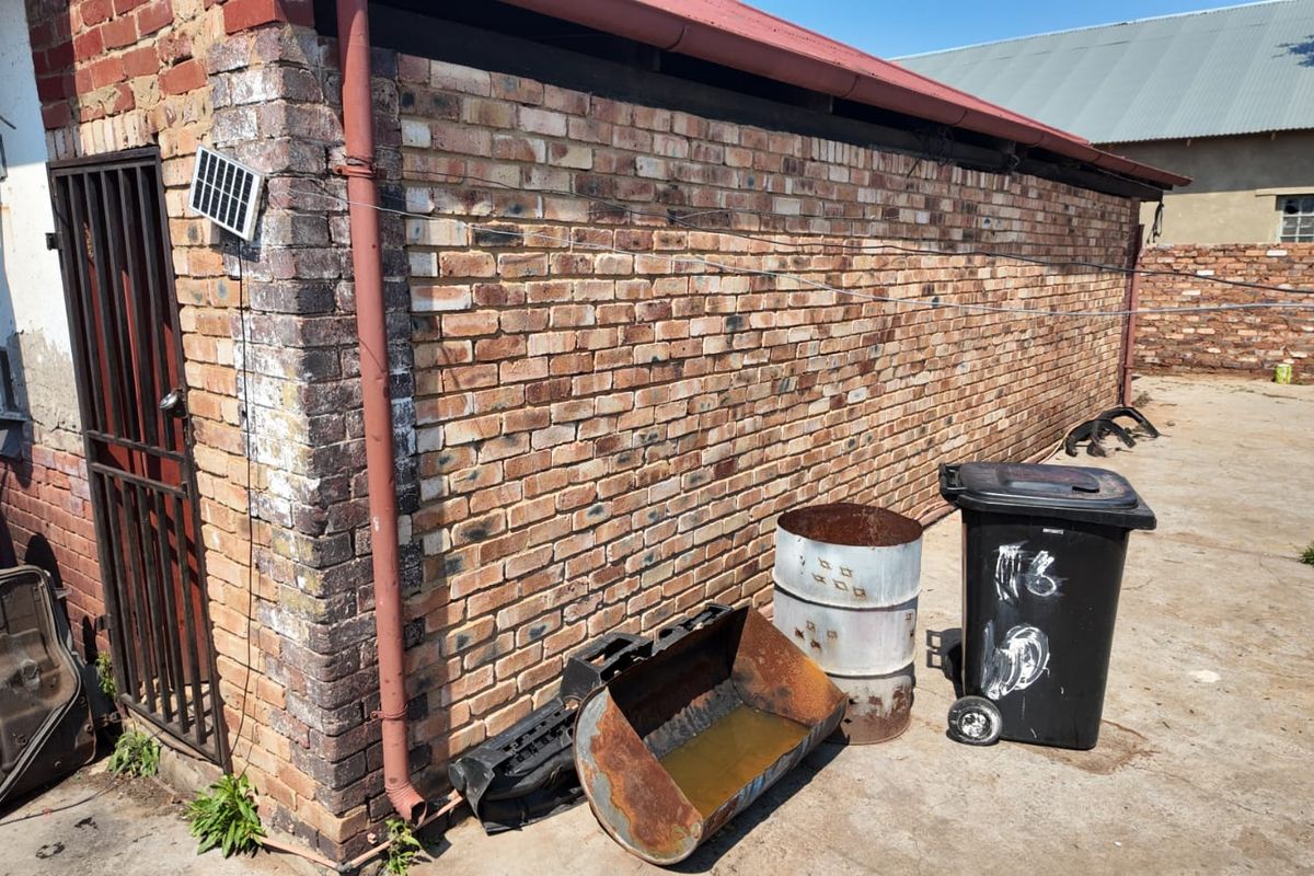 Rear exterior view of a brick building featuring a metal security gate, downpipe, and concrete paved area.