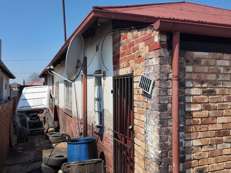 Rear exterior view of the property featuring exposed brickwork, a metal security gate, and corrugated roofing, showing signs of significant wear and maintenance requirements.