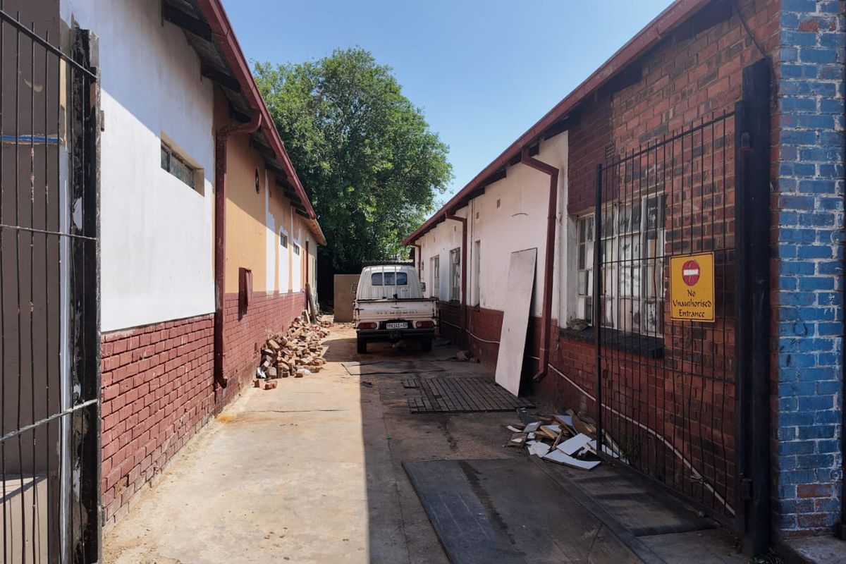 Rear exterior alleyway between industrial buildings featuring brick facades, concrete paving, and metal security fencing.