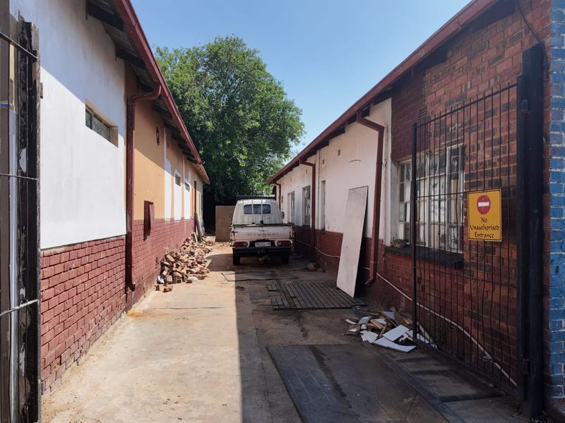 Rear exterior alleyway between industrial buildings featuring brick facades, concrete paving, and metal security fencing.