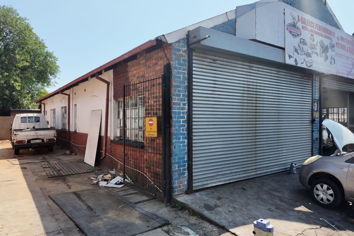 Front exterior view of a commercial workshop property featuring roller shutter doors, brick facade, and a concrete driveway area.