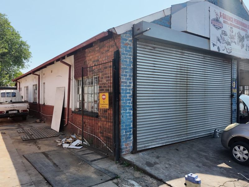 Front exterior view of a commercial workshop property featuring roller shutter doors, brick facade, and a concrete driveway area.