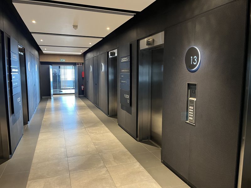 Modern office building elevator lobby featuring sleek metal wall paneling, tiled flooring, and recessed ceiling lighting on the 13th floor.