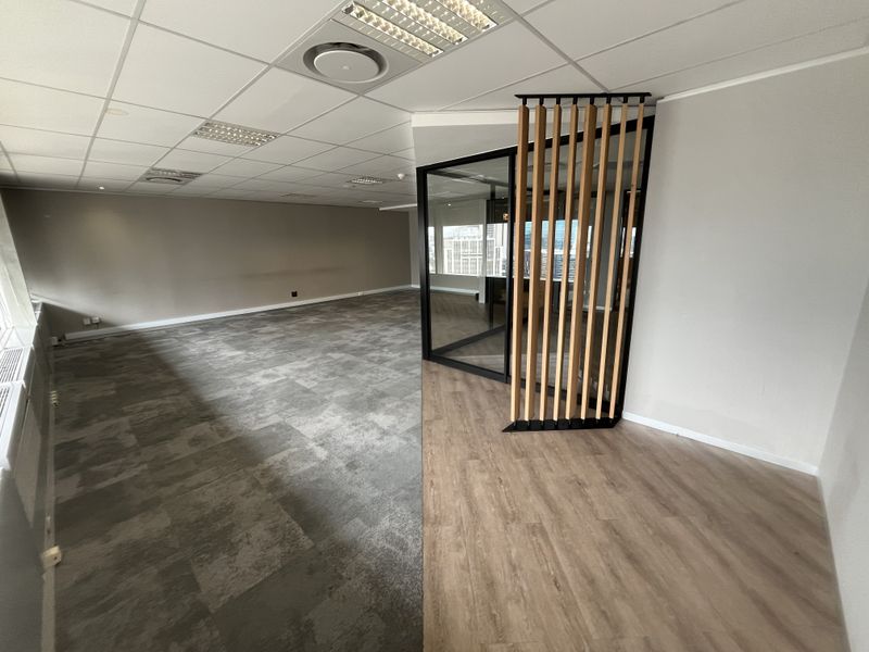 A modern, open-plan office space featuring a mix of carpet and laminate flooring, a glass-enclosed meeting area, and a decorative timber slat screen.