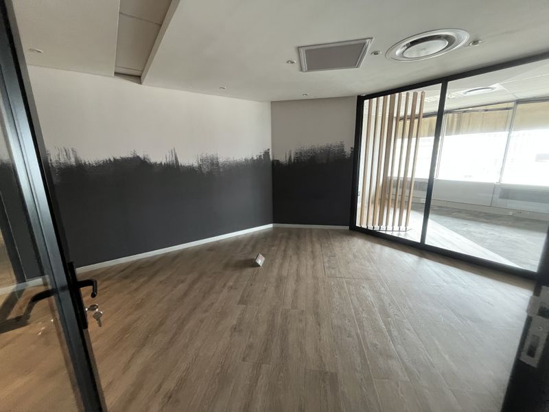A modern, empty office space featuring wood-look laminate flooring, a glass partition wall with timber slats, and recessed ceiling lighting.