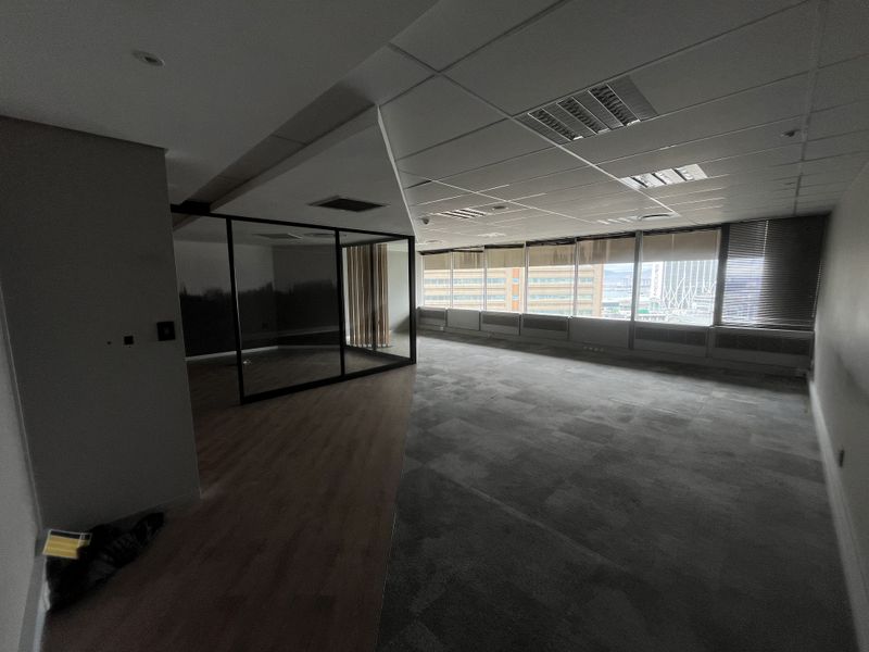 A spacious, vacant office suite featuring a mix of carpet and laminate flooring, a glass-partitioned meeting area, and large windows with city views.