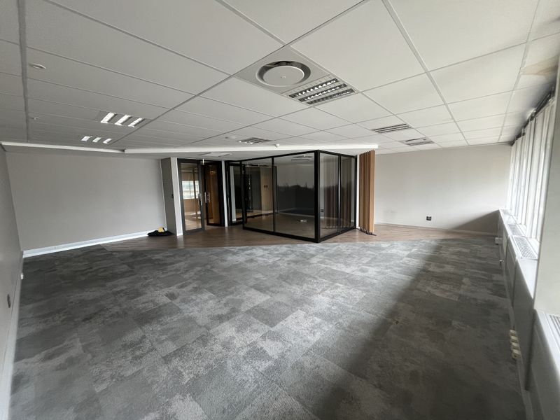 Spacious open-plan office area featuring a modern glass-enclosed meeting room, suspended ceilings, and a combination of carpet and laminate flooring.