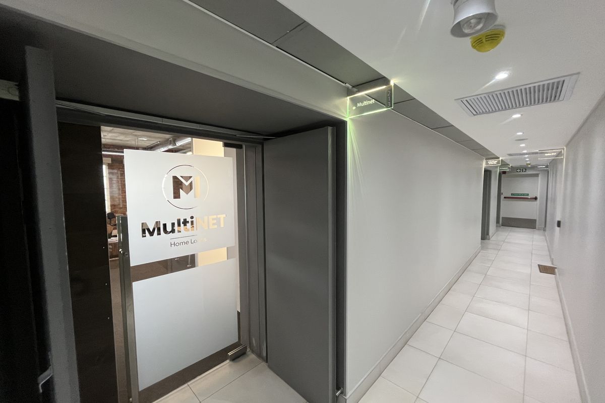 A clean, modern office hallway featuring tiled flooring, recessed lighting, and clear directional signage leading to the office entrance.