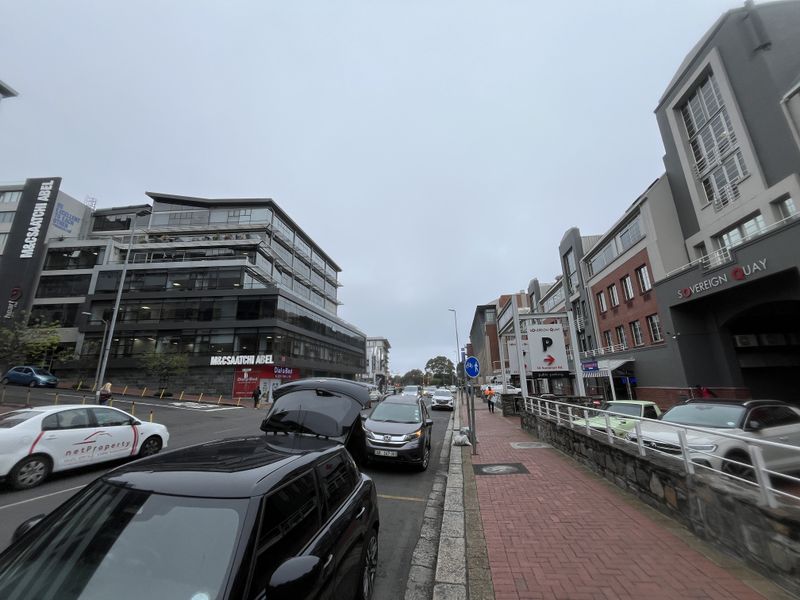 A street-level view of a commercial office district featuring modern building facades, paved pedestrian walkways, and public parking signage.