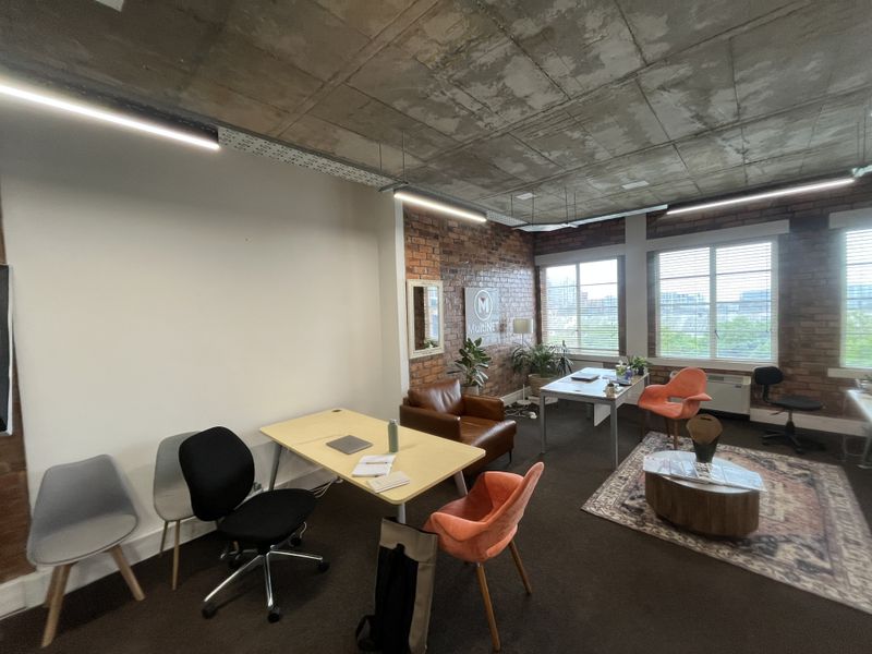 Modern open-plan office space featuring industrial-style exposed concrete ceilings, brick feature walls, and large windows providing ample natural light.