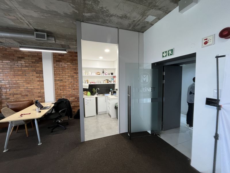 Modern office space featuring an exposed concrete ceiling, brick accent wall, and an integrated kitchenette, with a glass entrance door and professional fire safety fixtures.