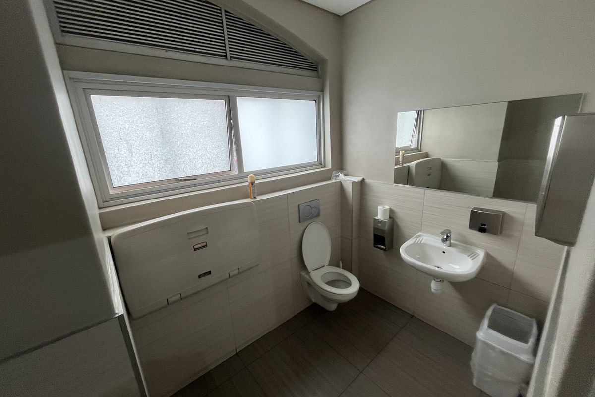A clean, modern commercial bathroom featuring a wall-mounted toilet, basin, and integrated baby changing station with neutral tiled finishes.