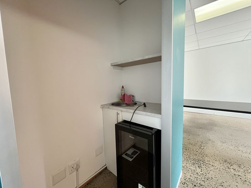A compact kitchenette or utility nook featuring a built-in sink, base cabinetry, and a wall-mounted shelf, situated within an office environment.