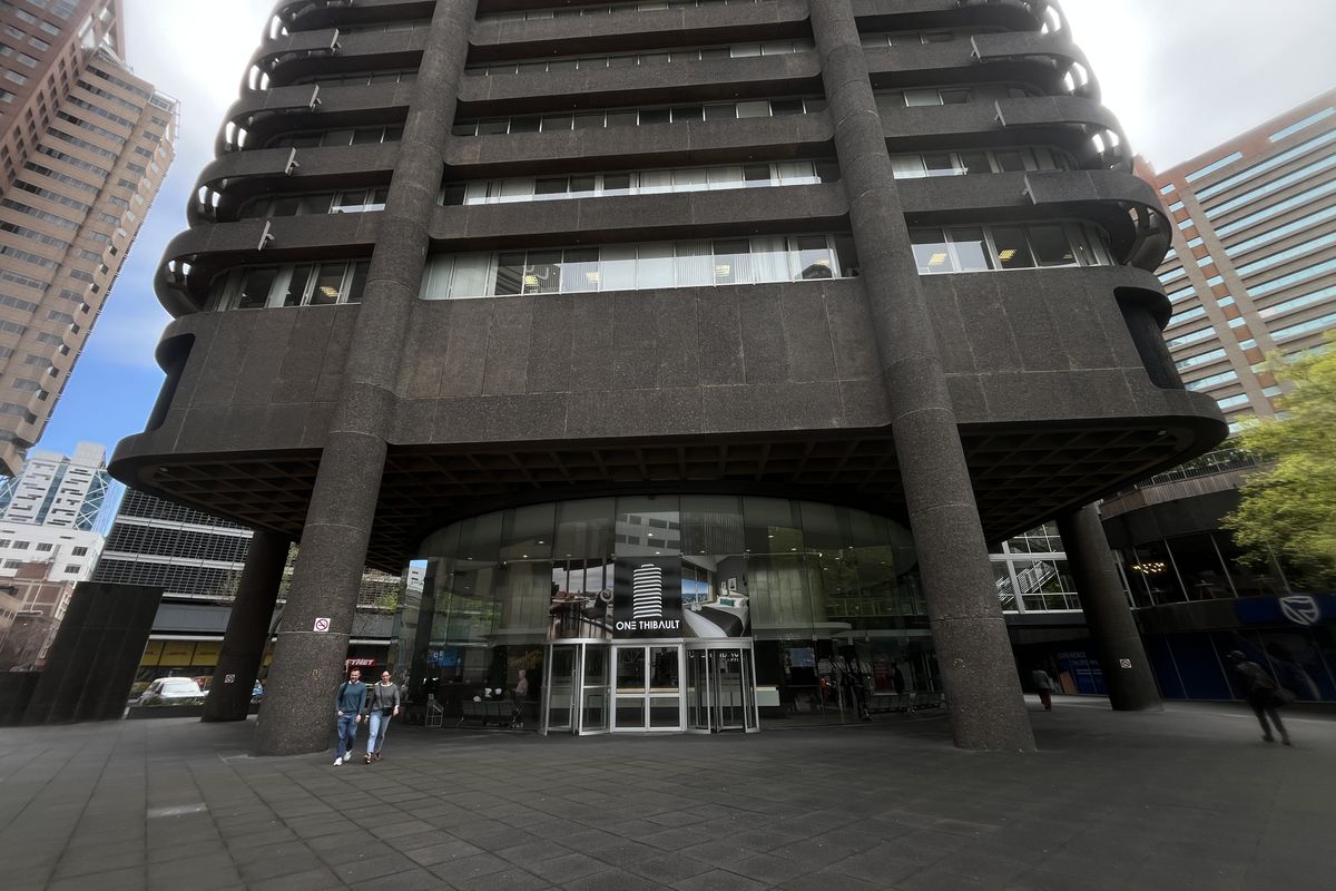 A low-angle view of the One Thibault office building entrance, featuring a prominent concrete facade, structural columns, and a modern glass revolving door.