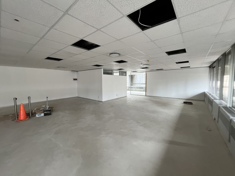 An open-plan office space featuring large windows, perimeter heating units, and a suspended ceiling with several missing tiles, awaiting final flooring installation.
