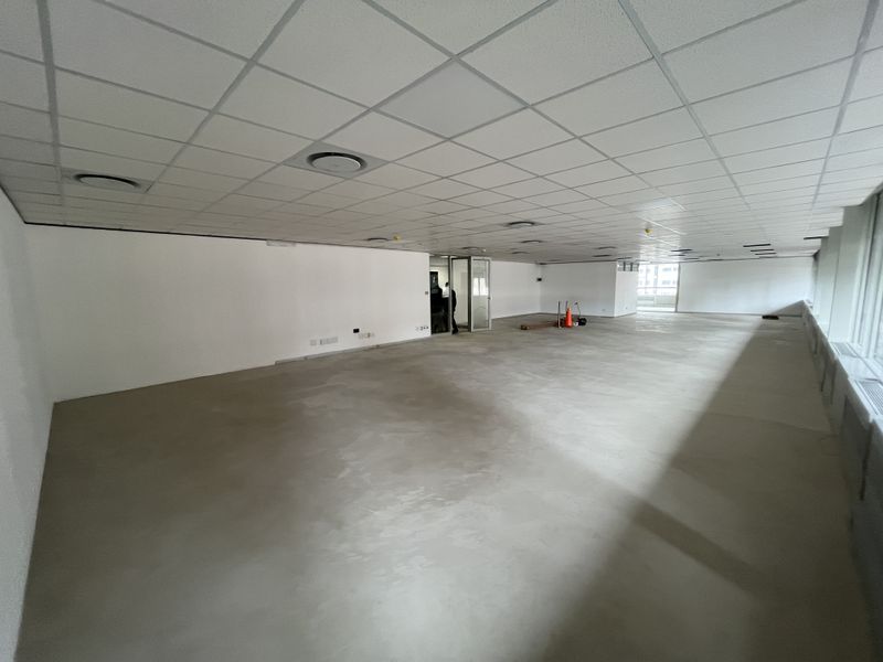 A spacious, open-plan office area featuring a suspended ceiling, recessed lighting, and large windows, currently presented as a blank canvas with unfinished flooring.