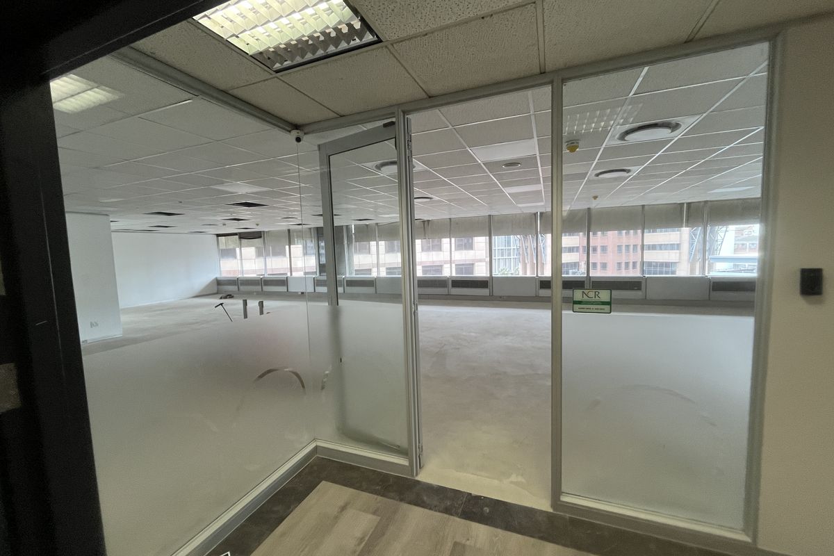 An empty, open-plan office space featuring glass partitions, suspended ceilings with recessed lighting, and large windows with blinds.