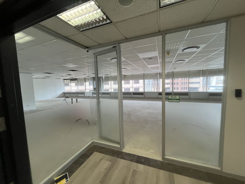 An empty, open-plan office space featuring glass partitions, suspended ceilings with recessed lighting, and large windows with blinds.
