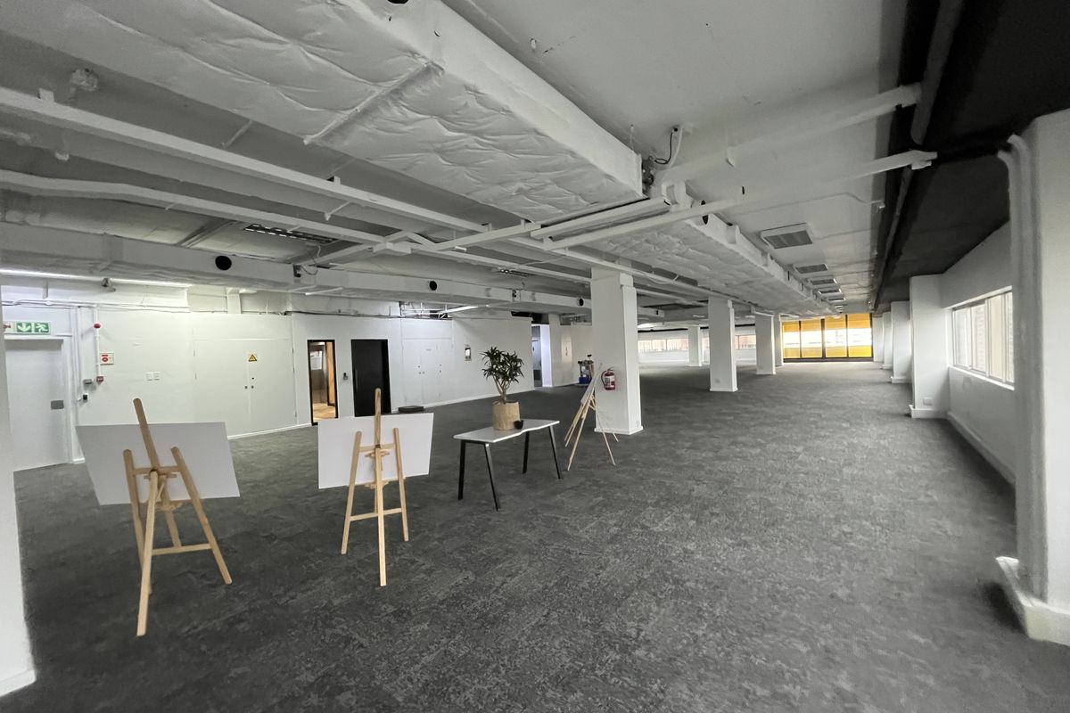 Spacious open-plan office area featuring exposed ceiling ducting, support columns, and neutral carpeted flooring, offering a versatile workspace layout.