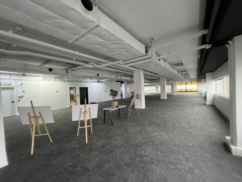 Spacious open-plan office area featuring exposed ceiling ducting, support columns, and neutral carpeted flooring, offering a versatile workspace layout.